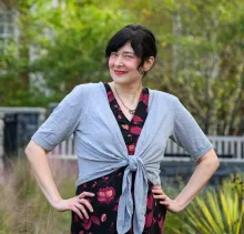 Librarian Emily Cook standing outdoors with hands on hips, smiling slightly. She has short dark hair and is wearing a black dress with a red floral pattern.